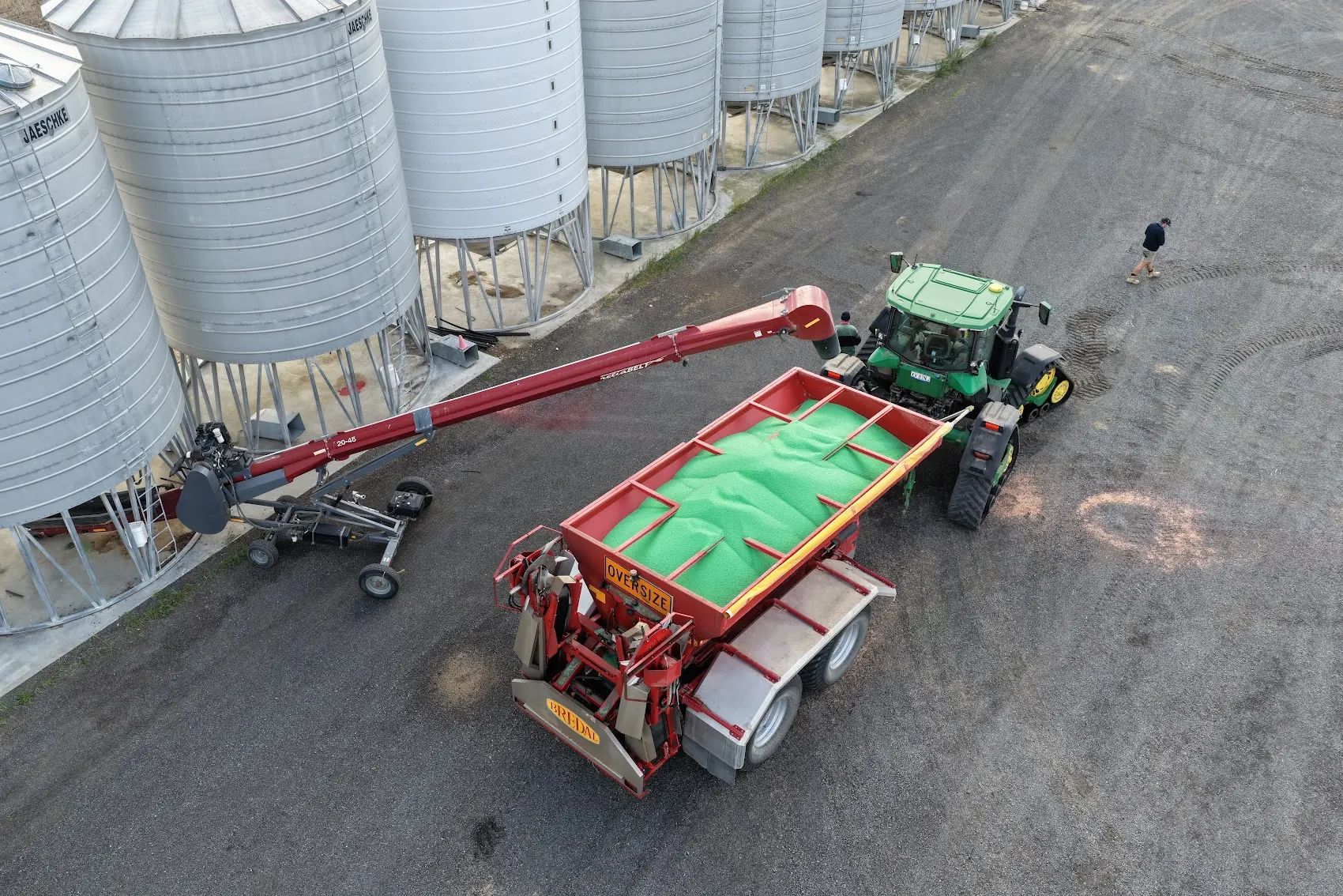 Aerial view of a red auger loading green N36 granules into a red spreader cart hitched to a green tractor, beside a row of grain silos, with a person standing near the tractor