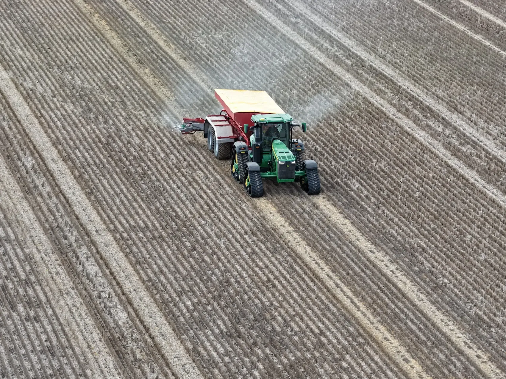 Aerial view of a green tractor towing a red spreader cart across rows of stubble, with fertiliser granules fanning out behind it